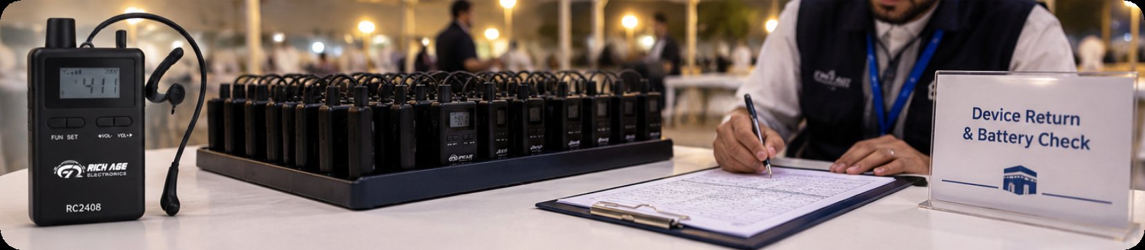 A coordinator logging receiver returns beside a Device Return and Battery Check sign, with a charging case of units on the desk A coordinator logging receiver returns beside a Device Return and Battery Check sign, with a charging case of units on the desk